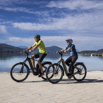 Two people riding e-bikes by a lake with mountains in the background