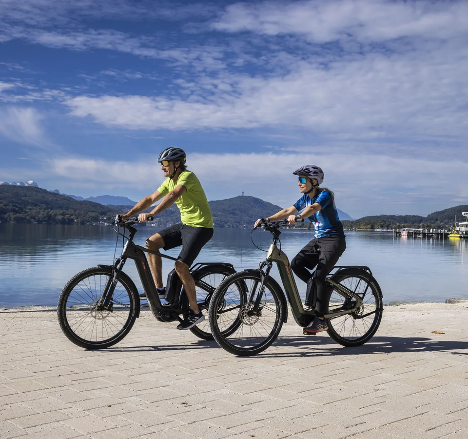 Klagenfurt kann mehr als Wörthersee! © Velontour - Heiko Mandl Zwei Personen fahren mit E-Bikes an einem See mit Bergen im Hintergrund