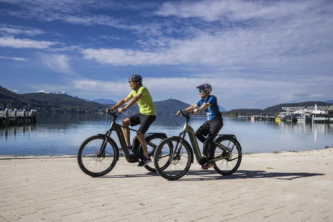 Zwei Personen fahren mit E-Bikes an einem See mit Bergen im Hintergrund