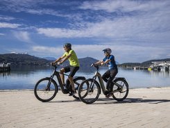 Two people riding e-bikes by a lake with mountains in the background