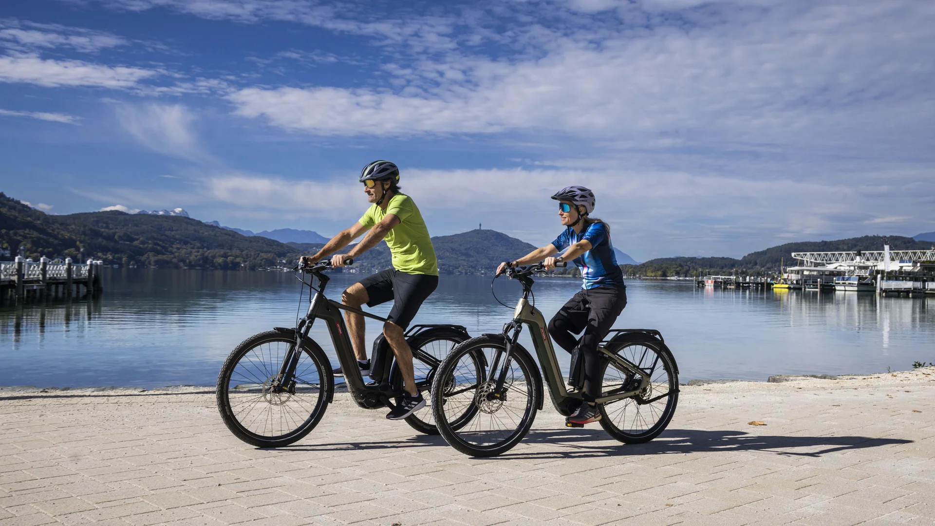 Zwei Personen fahren mit E-Bikes an einem See mit Bergen im Hintergrund