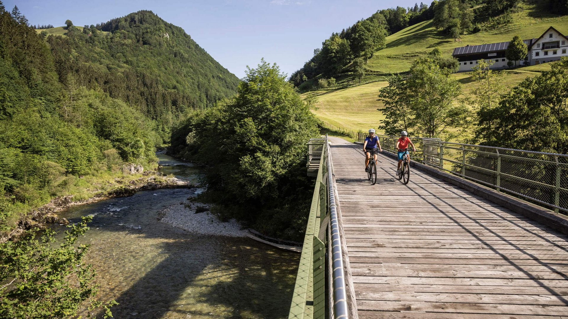 Two cyclists on a bridge over a river in a green mountain landscape