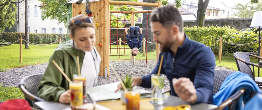 Parents sit in garden café as kids play on playground climbing structure