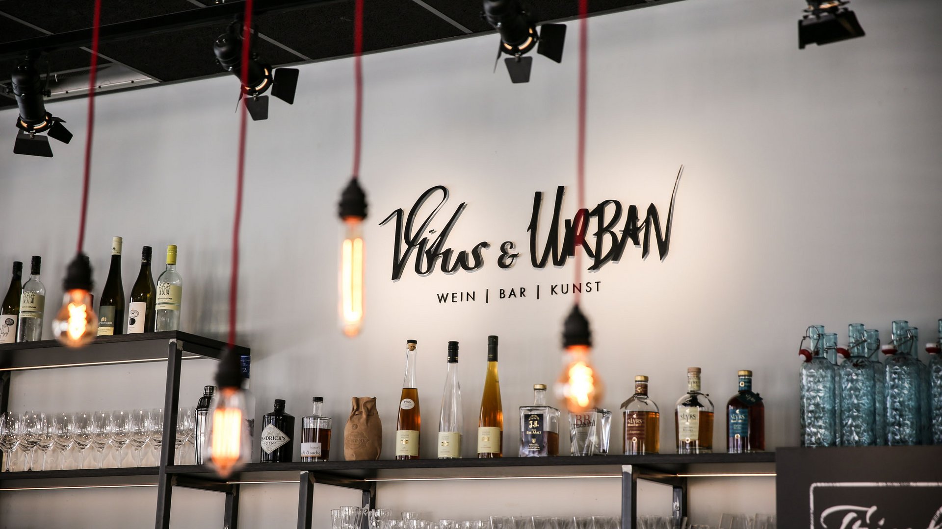 Bar shelves with bottles, glasses, and hanging lit light bulbs under logo