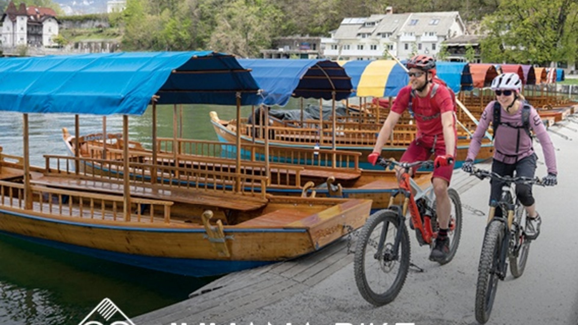Two cyclists riding bikes near wooden boats on a riverbank with forested hills