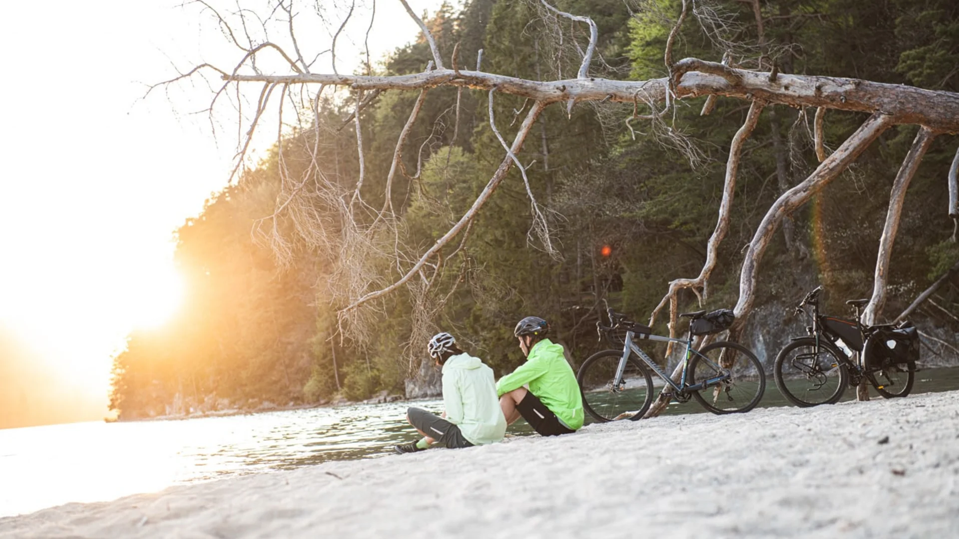 Two cyclists sitting on a beach at sunset next to their bikes