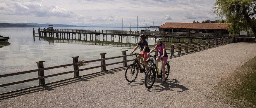 Zwei Radfahrer fahren auf einem Weg am See mit Steg und Bootshaus