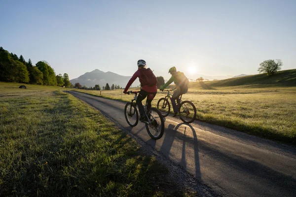Genussradeln mit Alpenblick im Tölzer Land © Tölzer Land Tourismus - Bernd Ritschel Zwei Radfahrer fahren auf Landstraße bei Sonnenaufgang