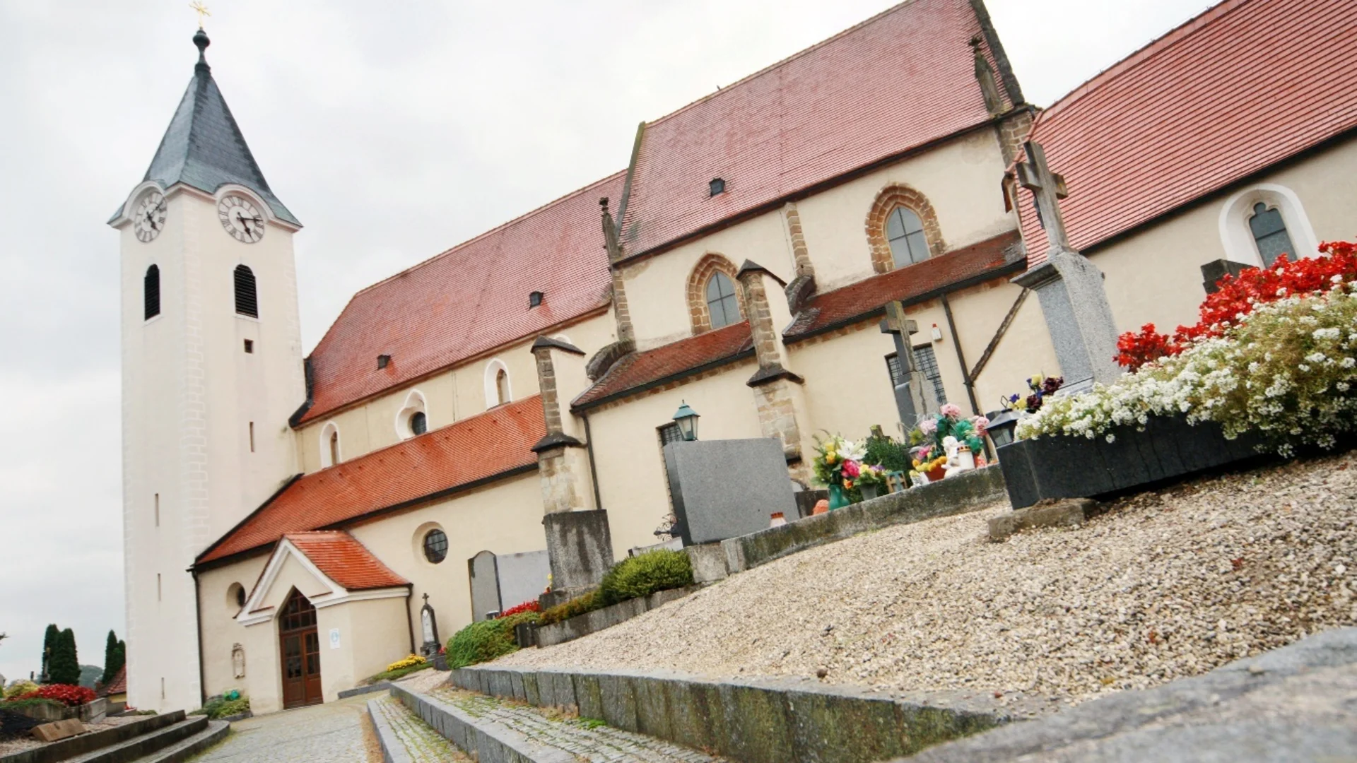 Tour-Detail Kirche mit rot gedecktem Dach und angrenzendem Friedhof mit Blumen