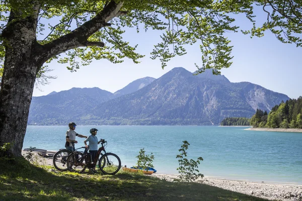Genussradeln mit Alpenblick im Tölzer Land © Velontour Zwei Radfahrer stehen unter einem Baum am See mit Blick auf Berge