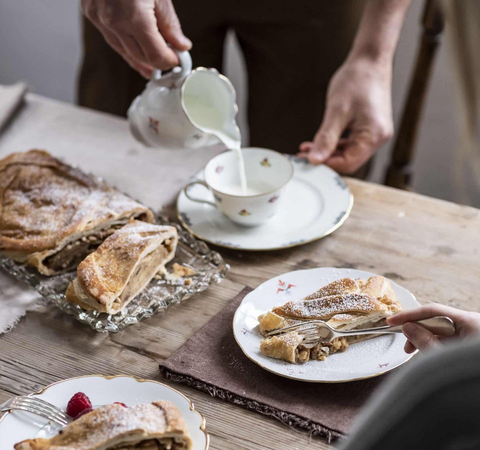 Person pouring milk into cup with apple strudel on wooden table