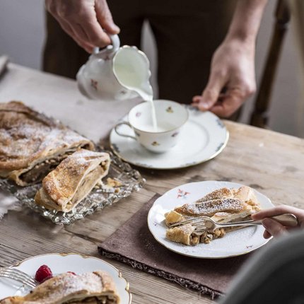 Person pouring milk into cup with apple strudel on wooden table
