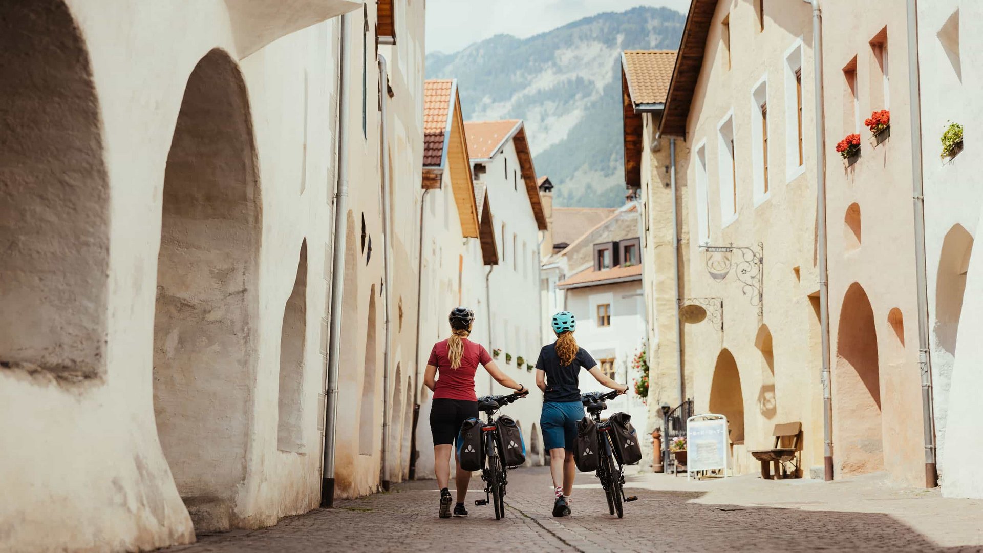 Two cyclists walking their bikes down a cobblestone street with mountain view