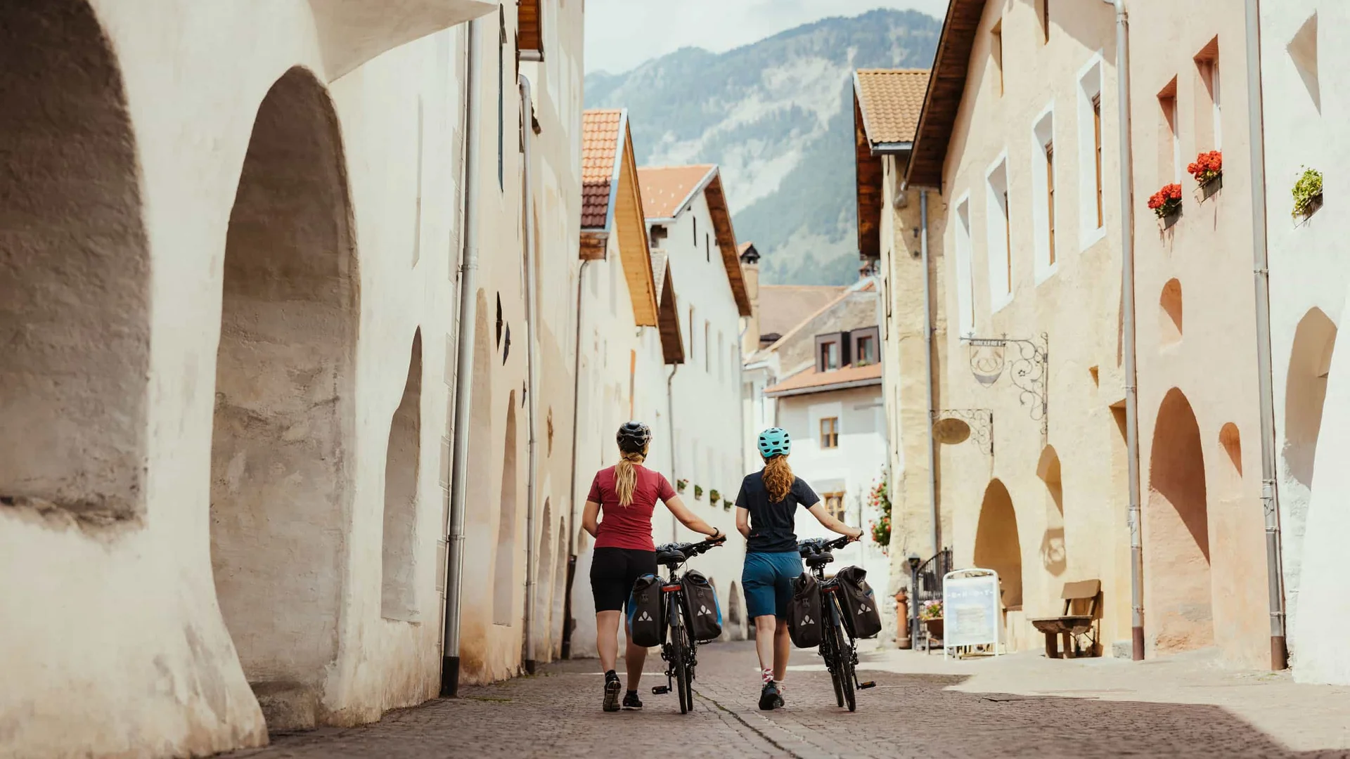 Zwei Radfahrer schieben ihre Fahrräder durch eine gepflasterte Straße mit Bergblick
