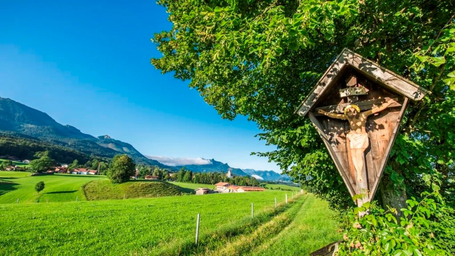 Holzkreuz mit Jesusfigur am Baum vor grüner Wiese und Bergen