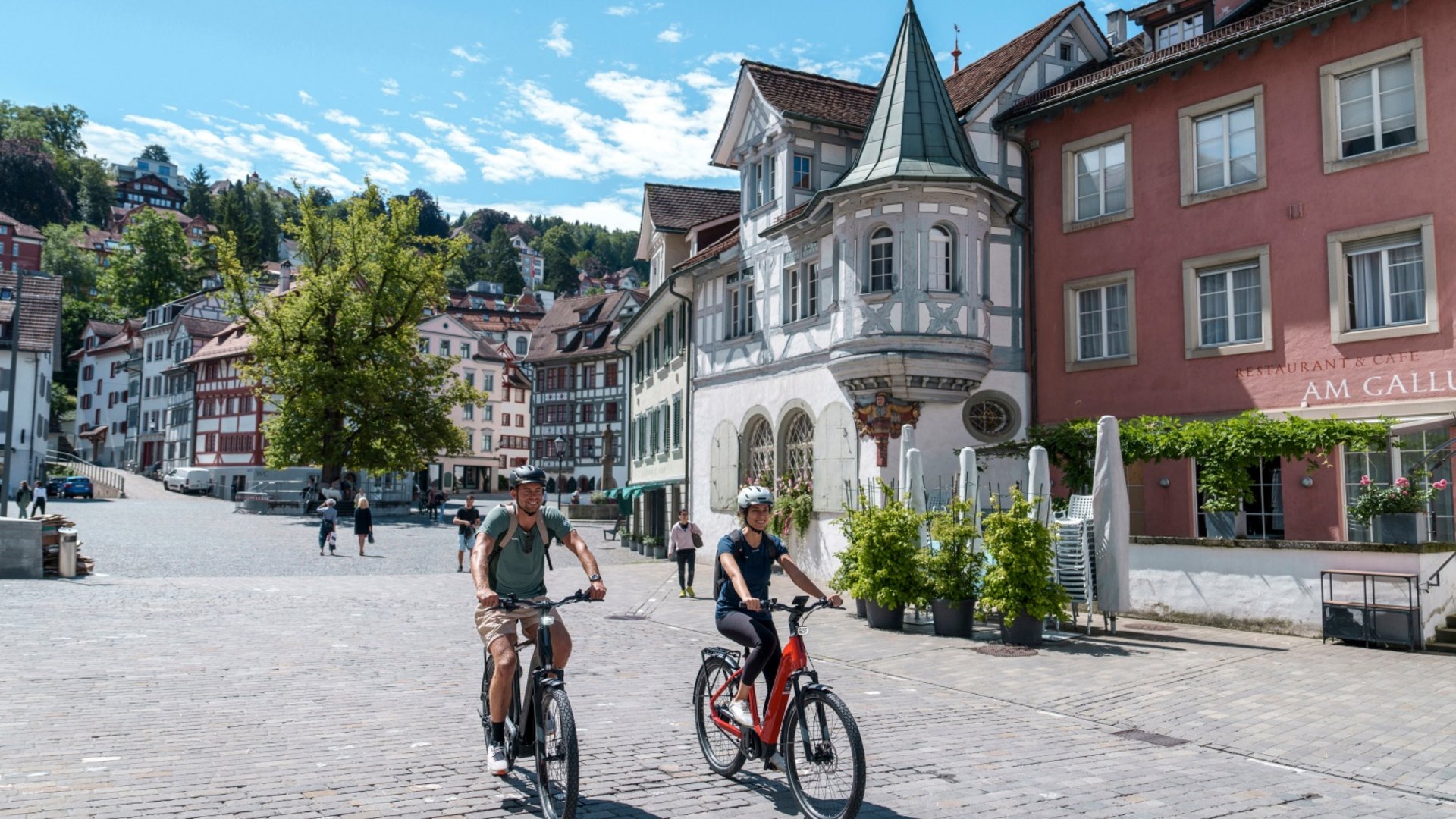Two people riding e-bikes through a historic town on a sunny day