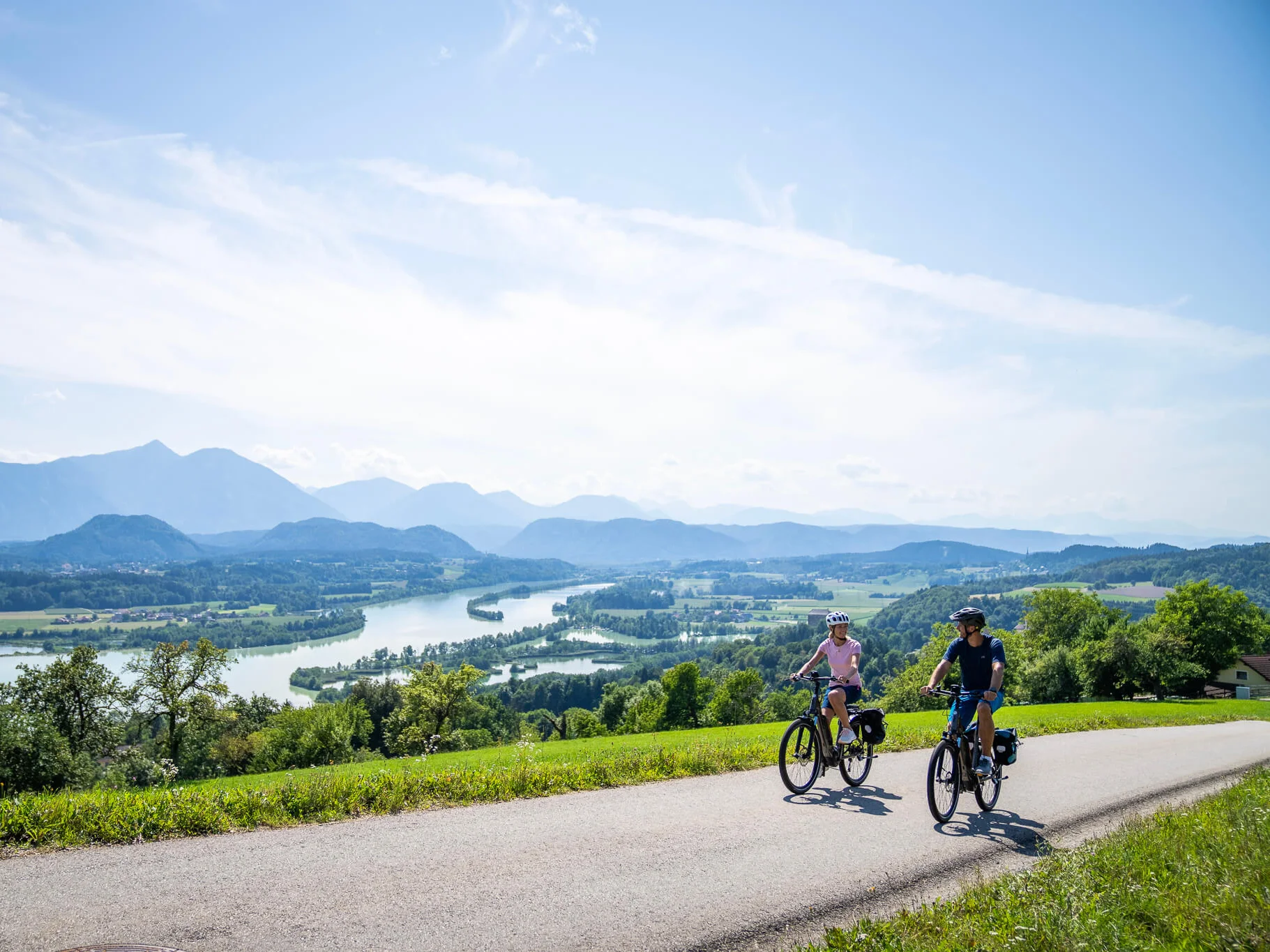 Kärnten © Kärnten Werbung - Gert Perauer Zwei Radfahrer fahren auf Landstraße mit Alpenblick und Fluss im Tal