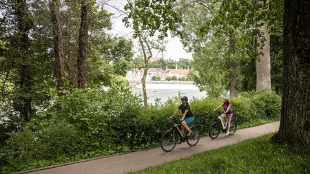 Zwei Personen fahren mit dem Fahrrad auf einem Waldweg am Flussufer
