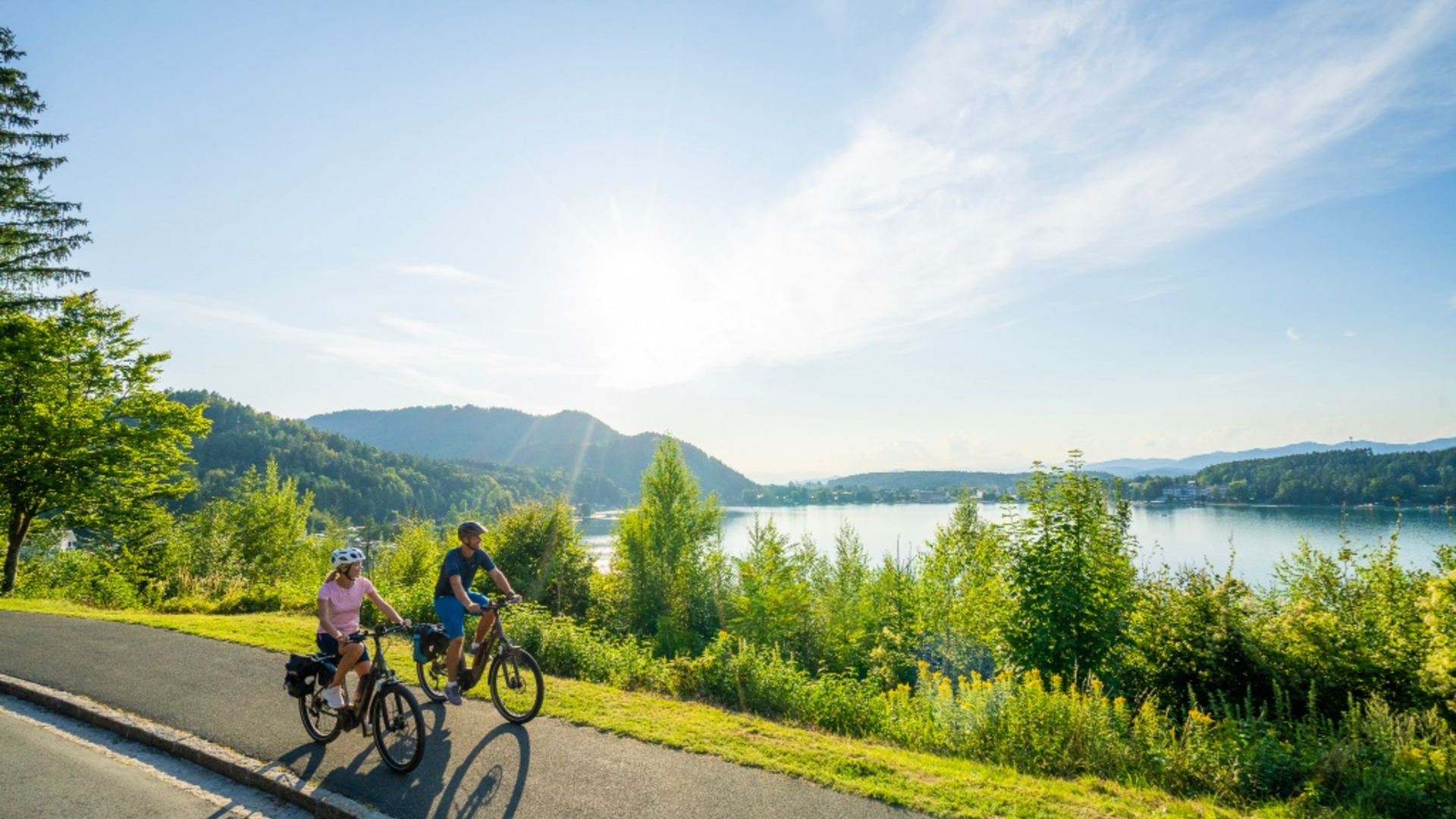 Two cyclists riding on a path beside a lake on a sunny day