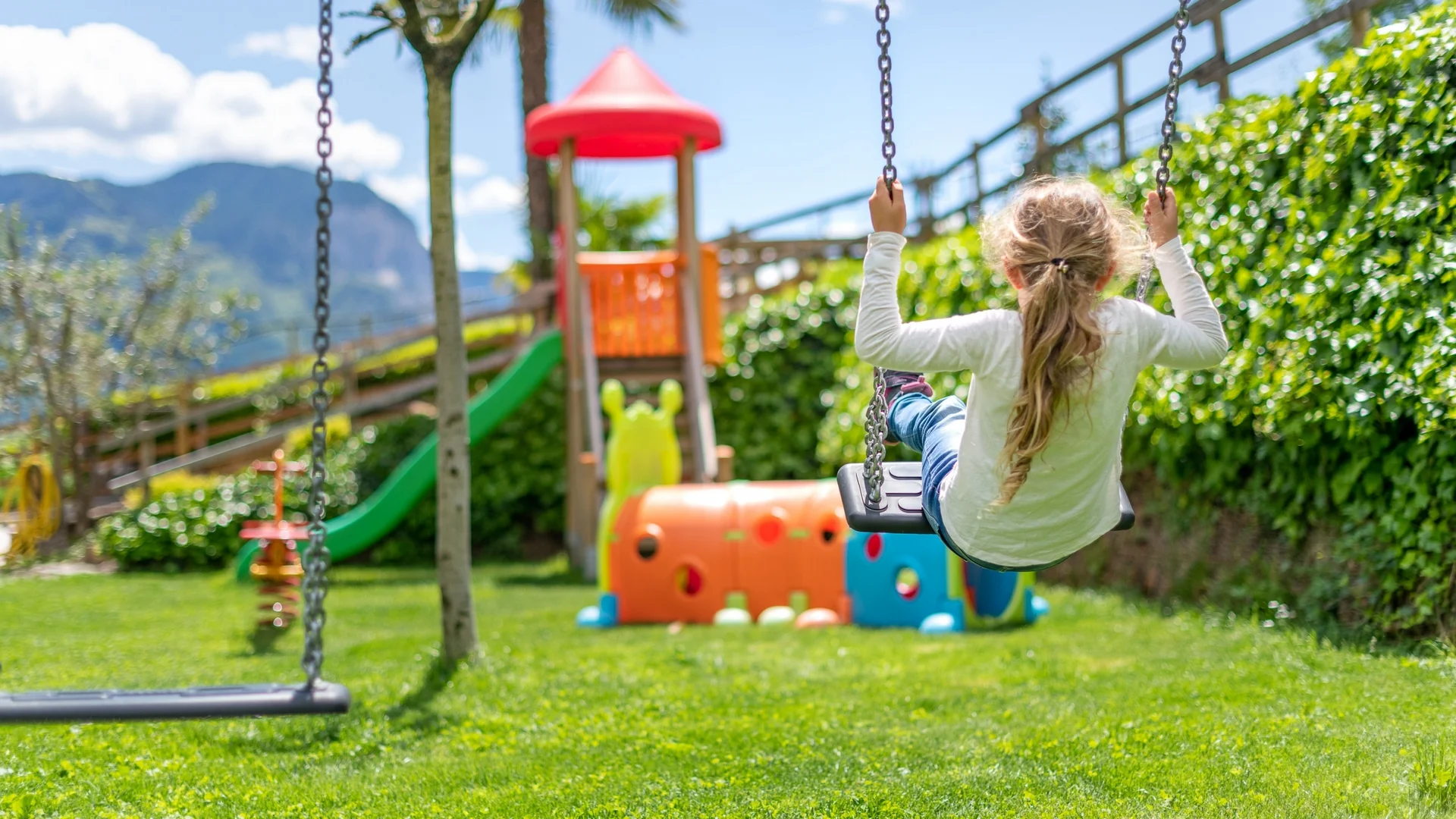 Mädchen schaukelt auf Spielplatz mit Rutsche und grünem Gras