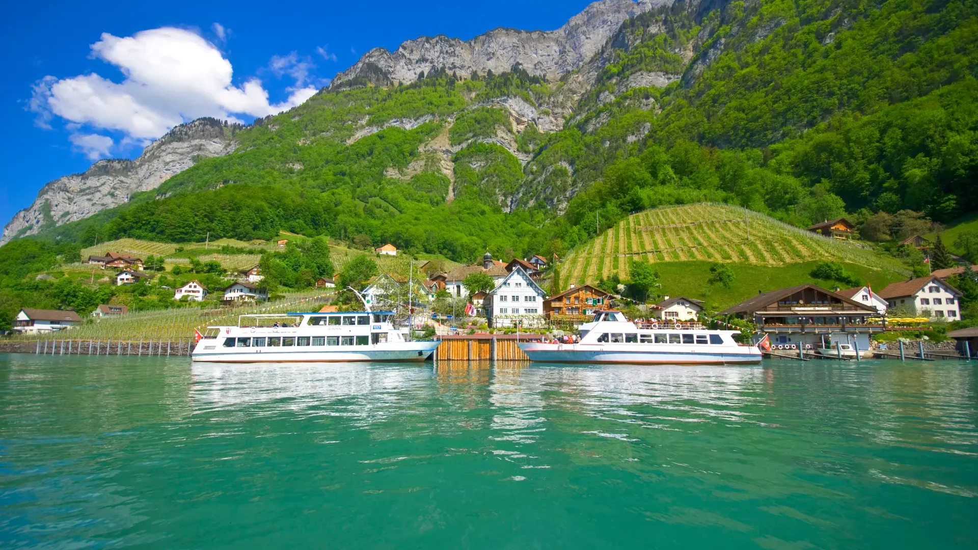 Boote auf See vor kleinem Dorf und bewaldetem Berg unter blauem Himmel
