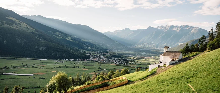 Bergtal mit Kirche auf grünem Hügel und Sonnenstrahlen durch Wolken