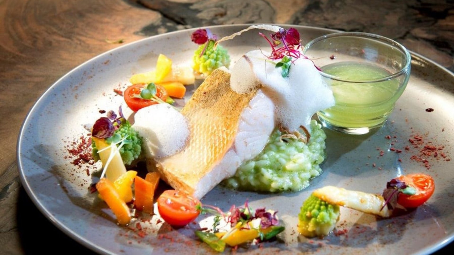 Gourmet dish with fish fillet, vegetables, foam, and green dip in a glass