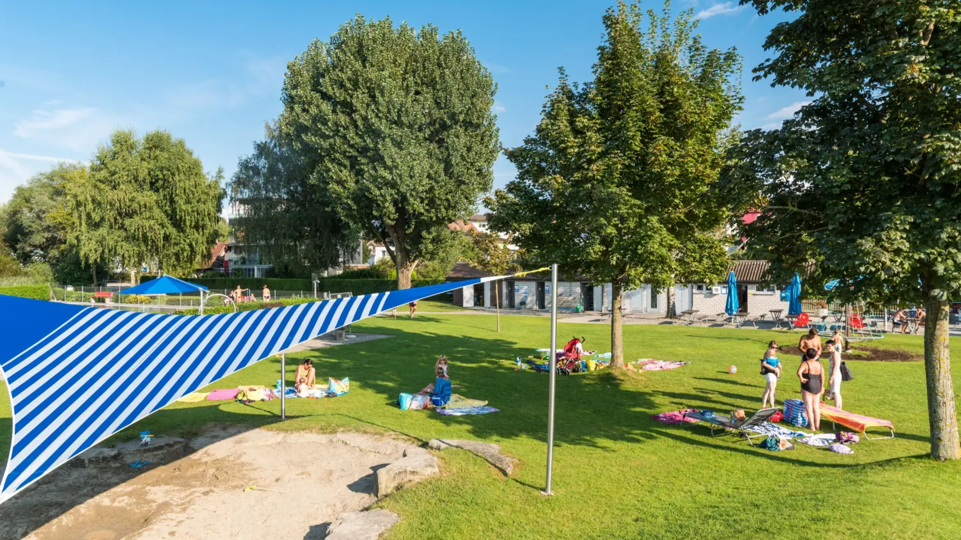 People relaxing in a park with large trees and sunshades on a sunny day