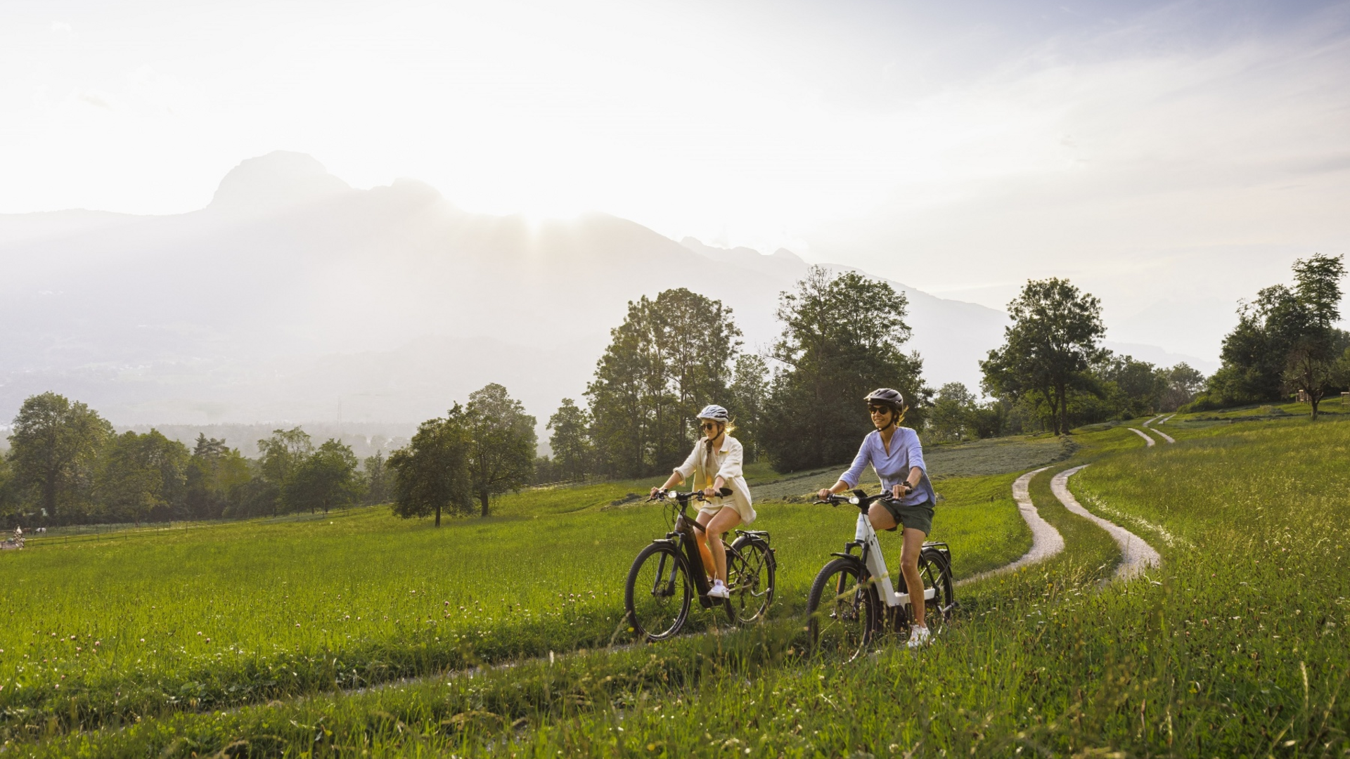 Two women cycling on a dirt path through a grassy field at sunset
