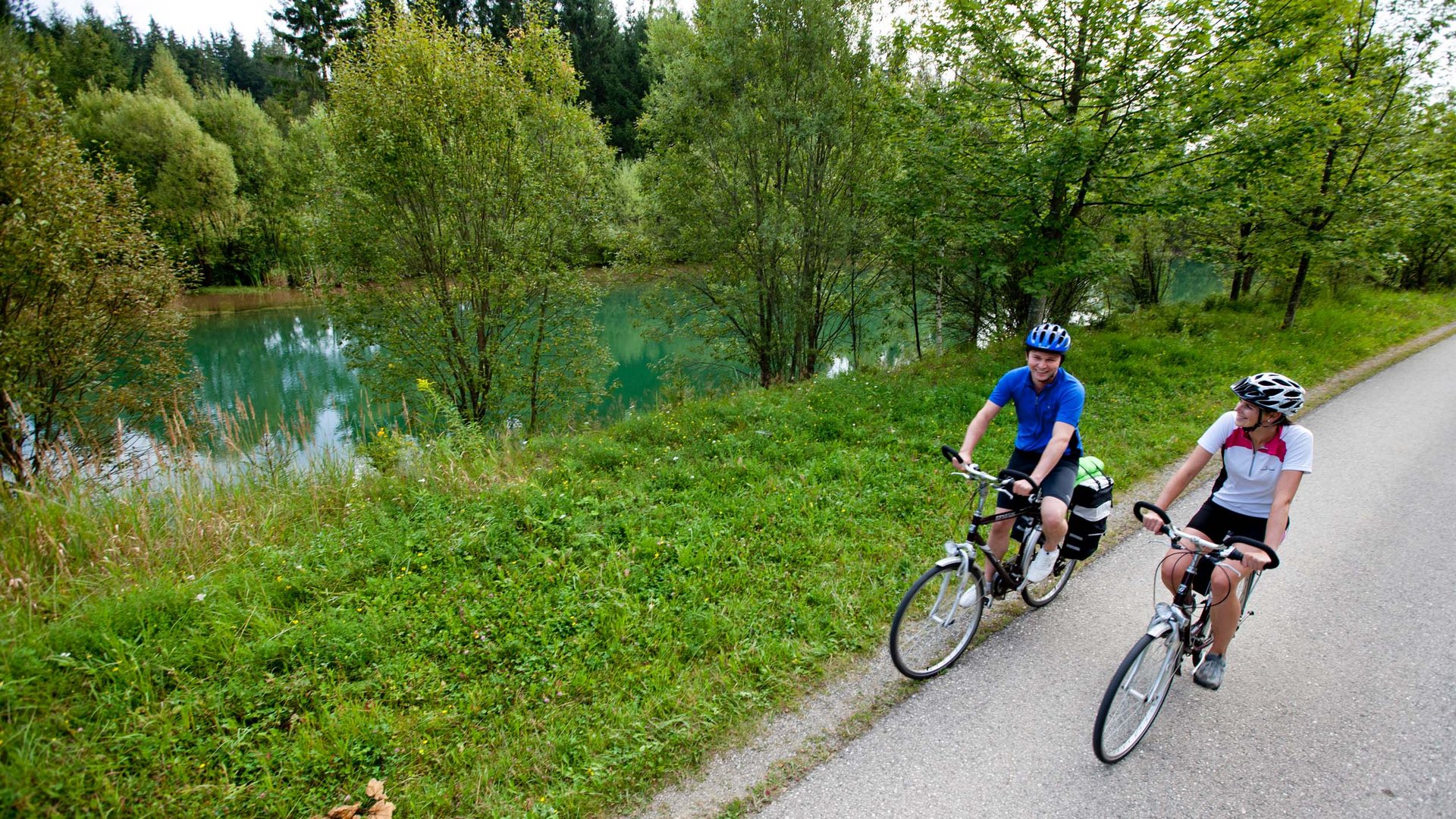 Two cyclists riding on a path beside a river and trees