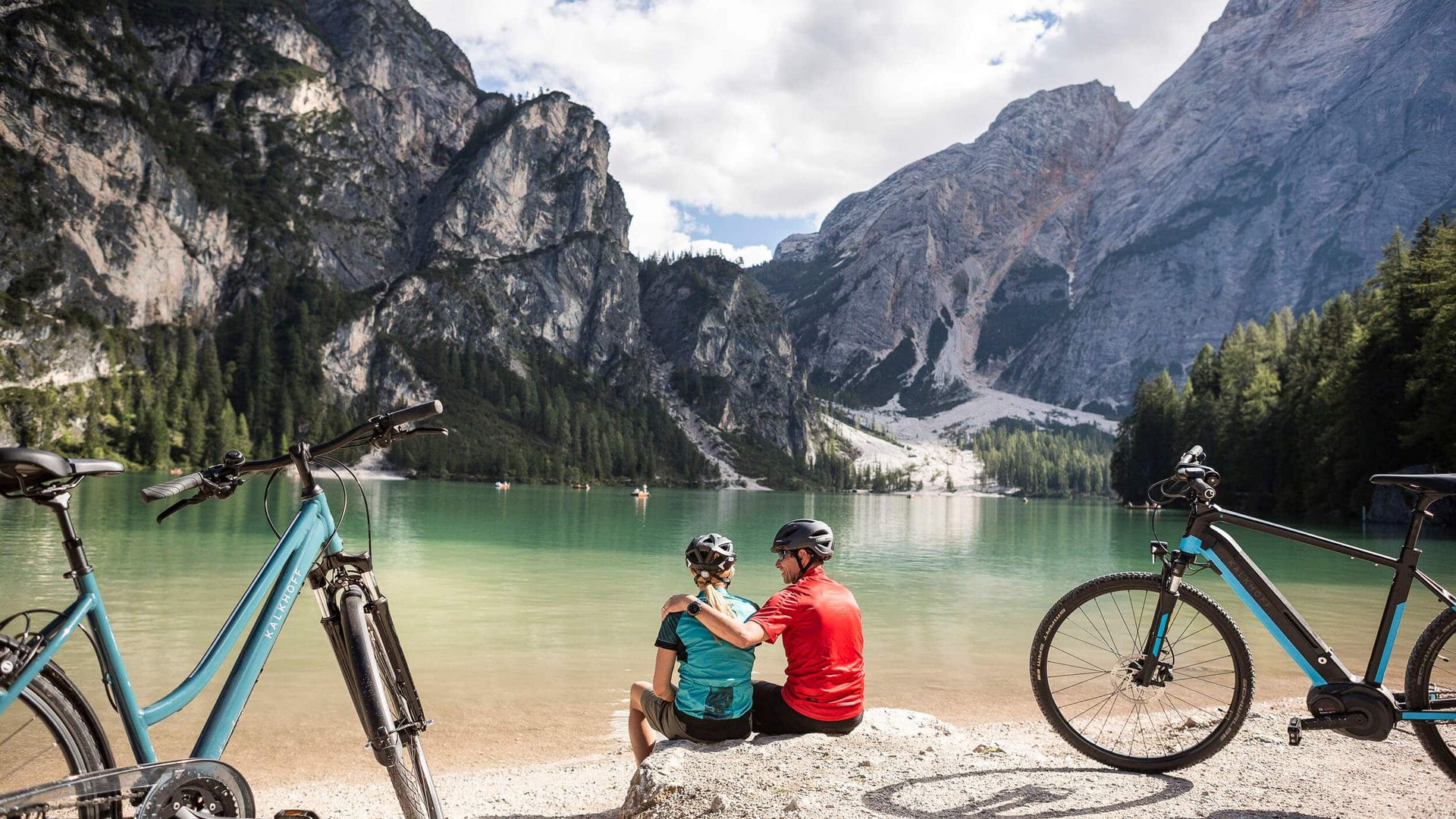 Couple with bikes sitting by lake surrounded by mountains on a sunny day