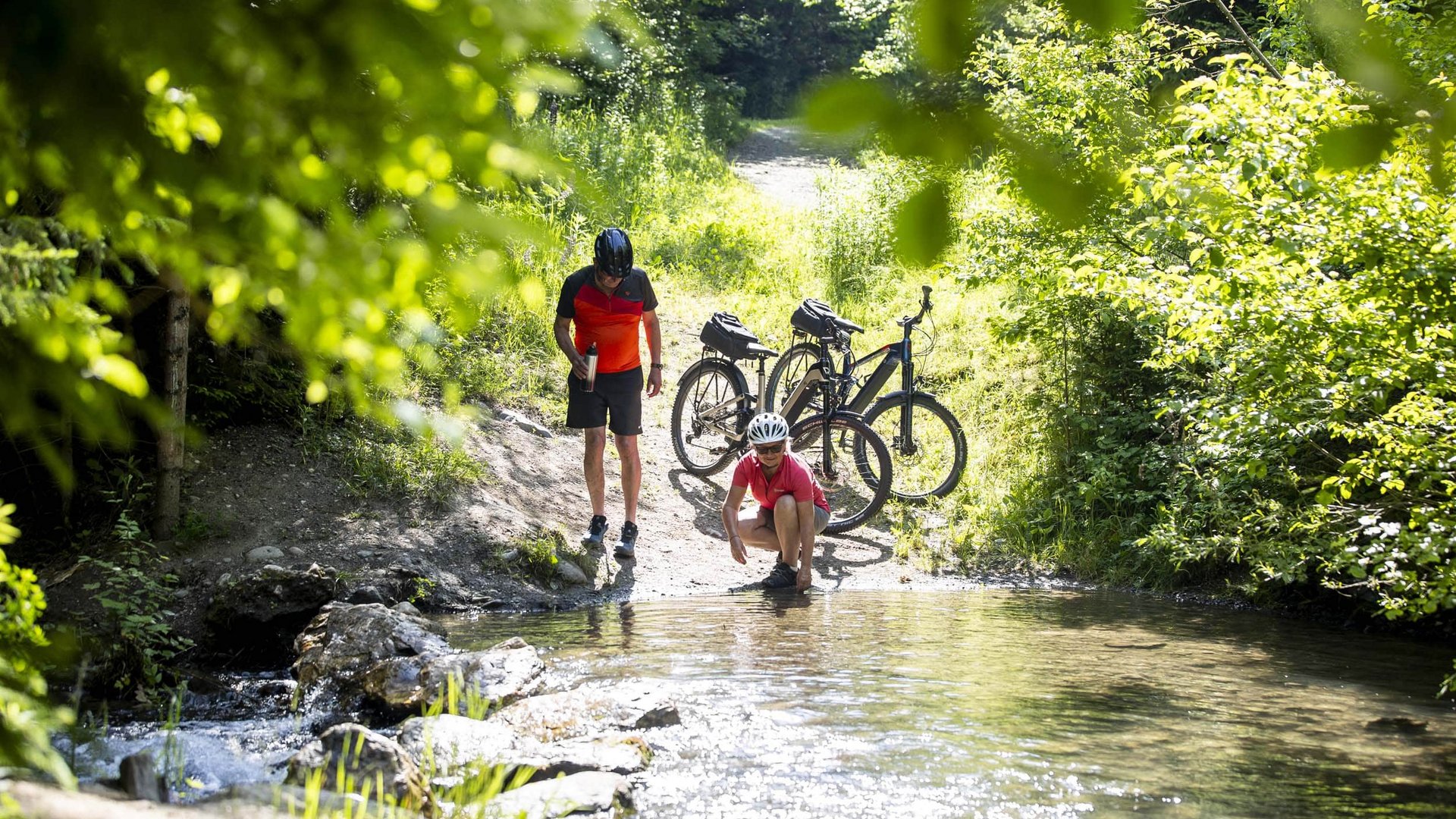Two cyclists taking a break by a stream in a sunny forest