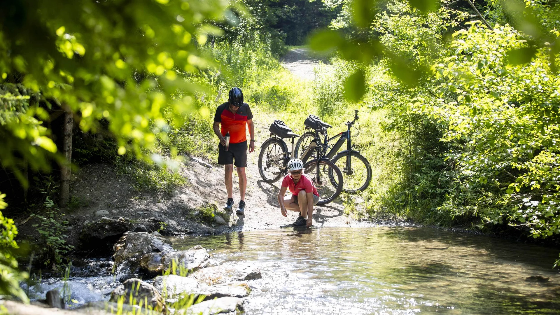Twee fietsers pauzeren bij een beek in een zonnig bos