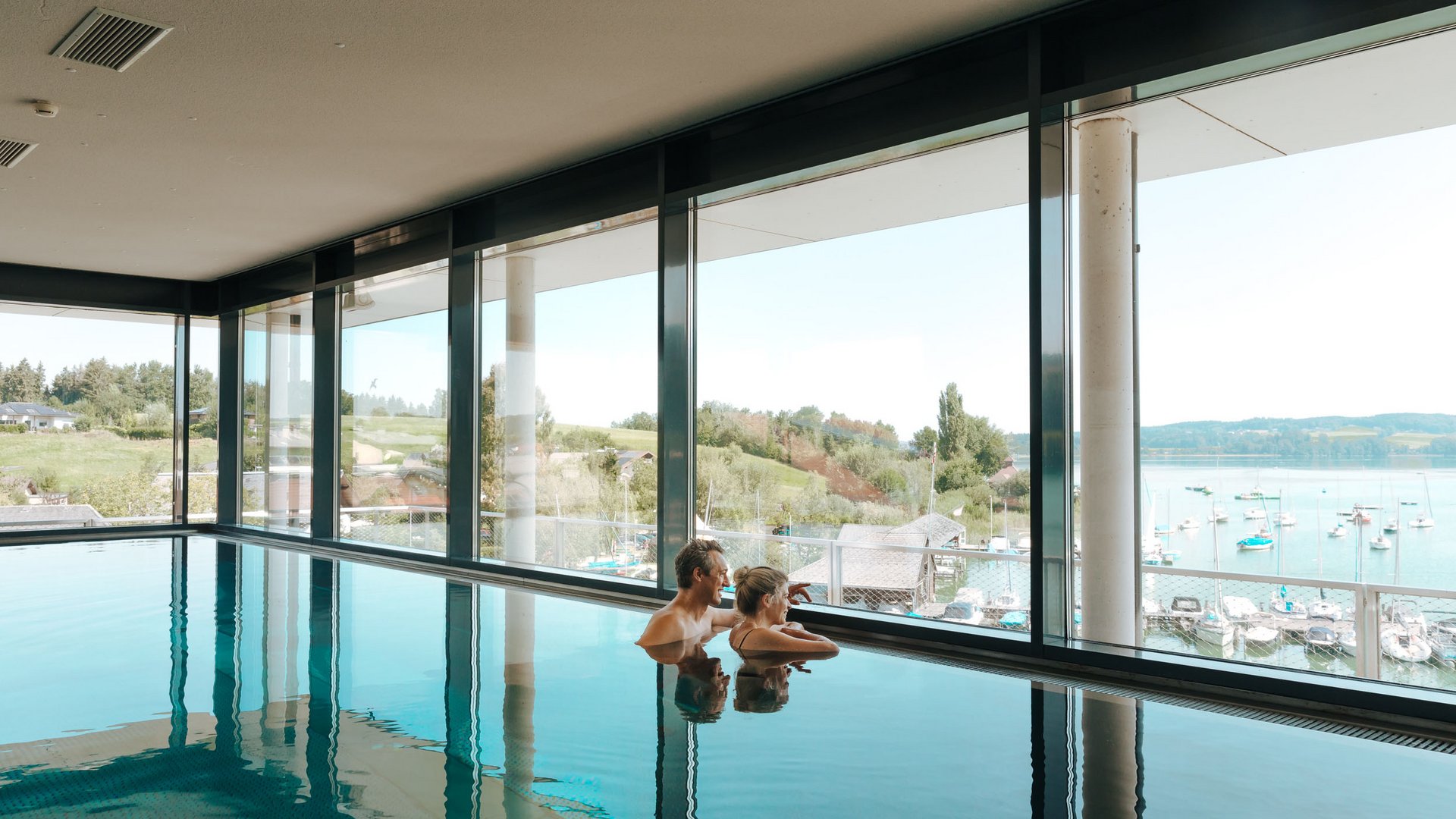 Couple in indoor pool looking out at lake and landscape through large windows