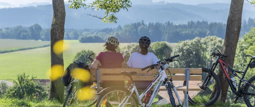 Two cyclists sitting on a bench overlooking green fields and mountains