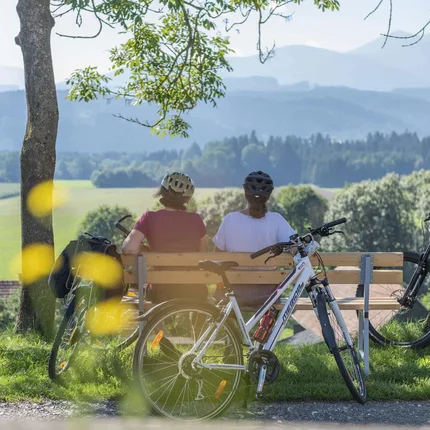 Two cyclists sitting on a bench overlooking green fields and mountains