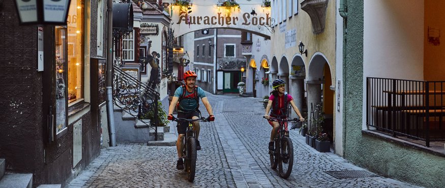 Two cyclists riding through a narrow, historic cobblestone street