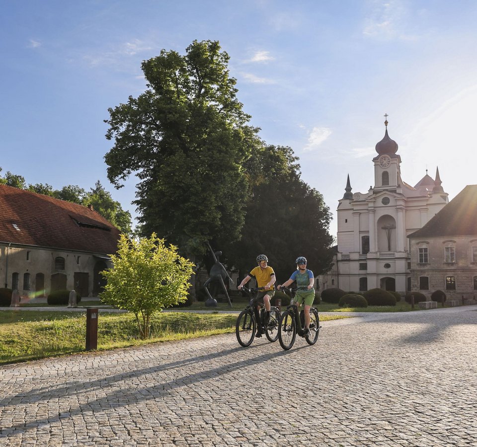 Zwei Radfahrer bei sonnigem Wetter vor historischen Gebäuden und Bäumen