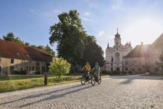Zwei Radfahrer bei sonnigem Wetter vor historischen Gebäuden und Bäumen