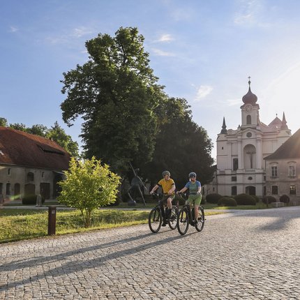 Zwei Radfahrer bei sonnigem Wetter vor historischen Gebäuden und Bäumen