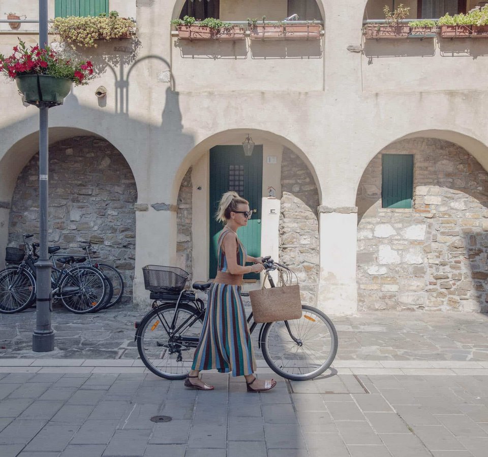 Frau schiebt Fahrrad auf gepflastertem Weg vor Gebäude mit Bögen
