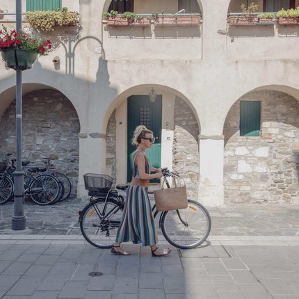 Frau schiebt Fahrrad auf gepflastertem Weg vor Gebäude mit Bögen
