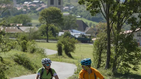 Drei Länder, du und dein Rad: Den Reschensee grenzüberschreitend entdecken © IDM Südtirol - T. Grüner Zwei Radfahrer fahren auf einem Weg in einer grünen ländlichen Gegend mit Kirche im Hintergrund.