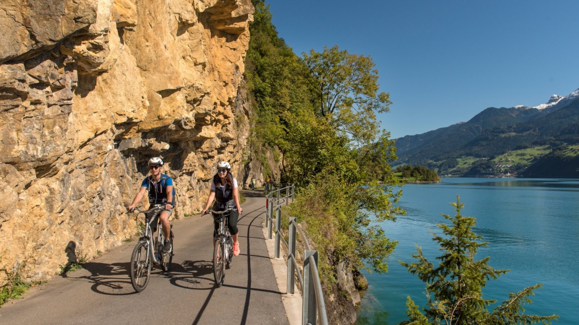Two cyclists on mountain path beside lake with cliffs and trees