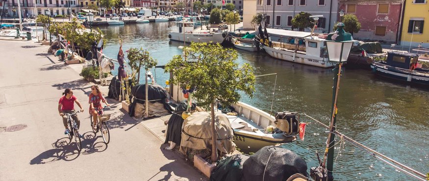 Two cyclists on a sunny canal with boats and colorful houses