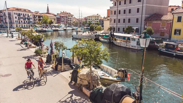 Grado: Herbst-Radeln an der Adria © Ivan Regolin Zwei Radfahrer am sonnigen Kanal mit Booten und bunten Häusern