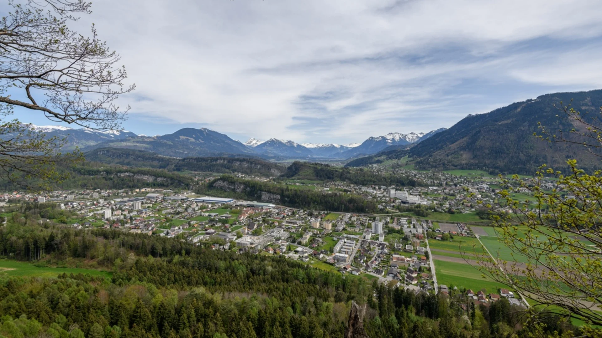 Blick auf eine Stadt in einem Tal mit bewaldeten Hügeln und schneebedeckten Bergen im Hintergrund