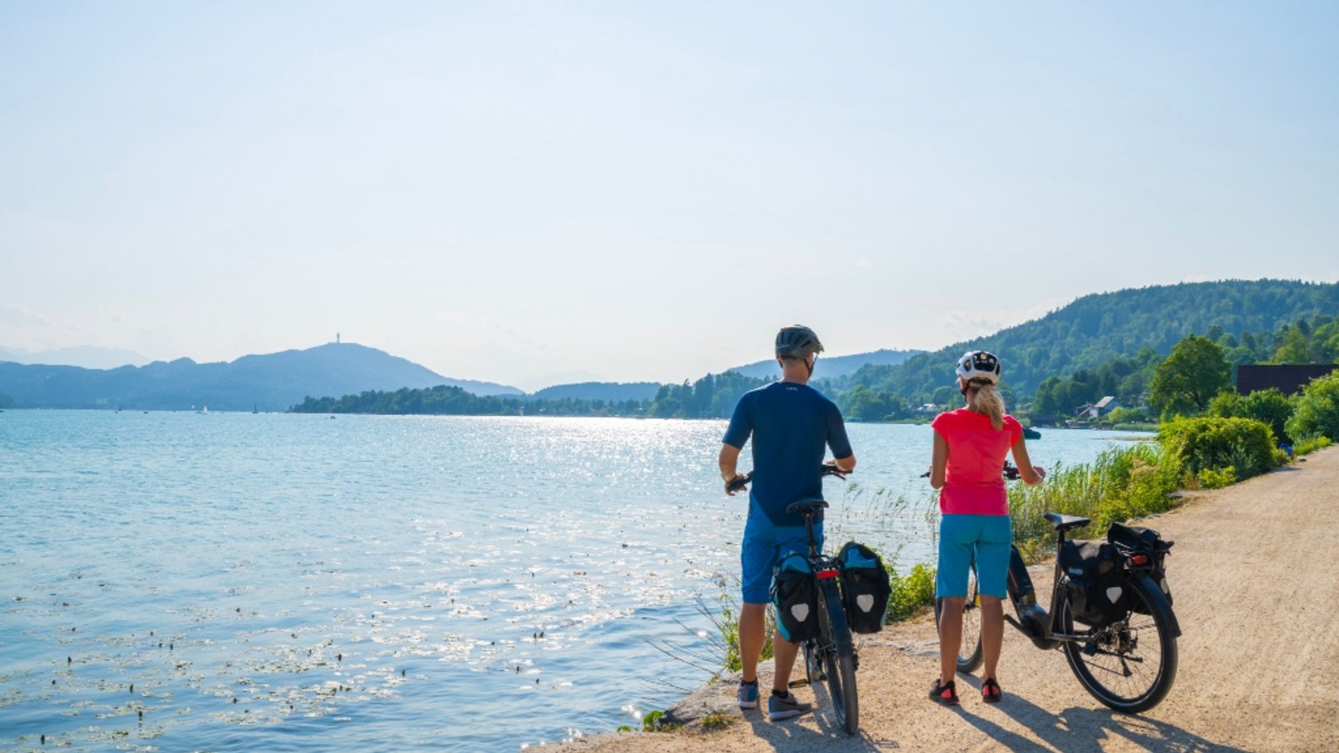 Two cyclists standing by a lakeside with mountains in the sunny background