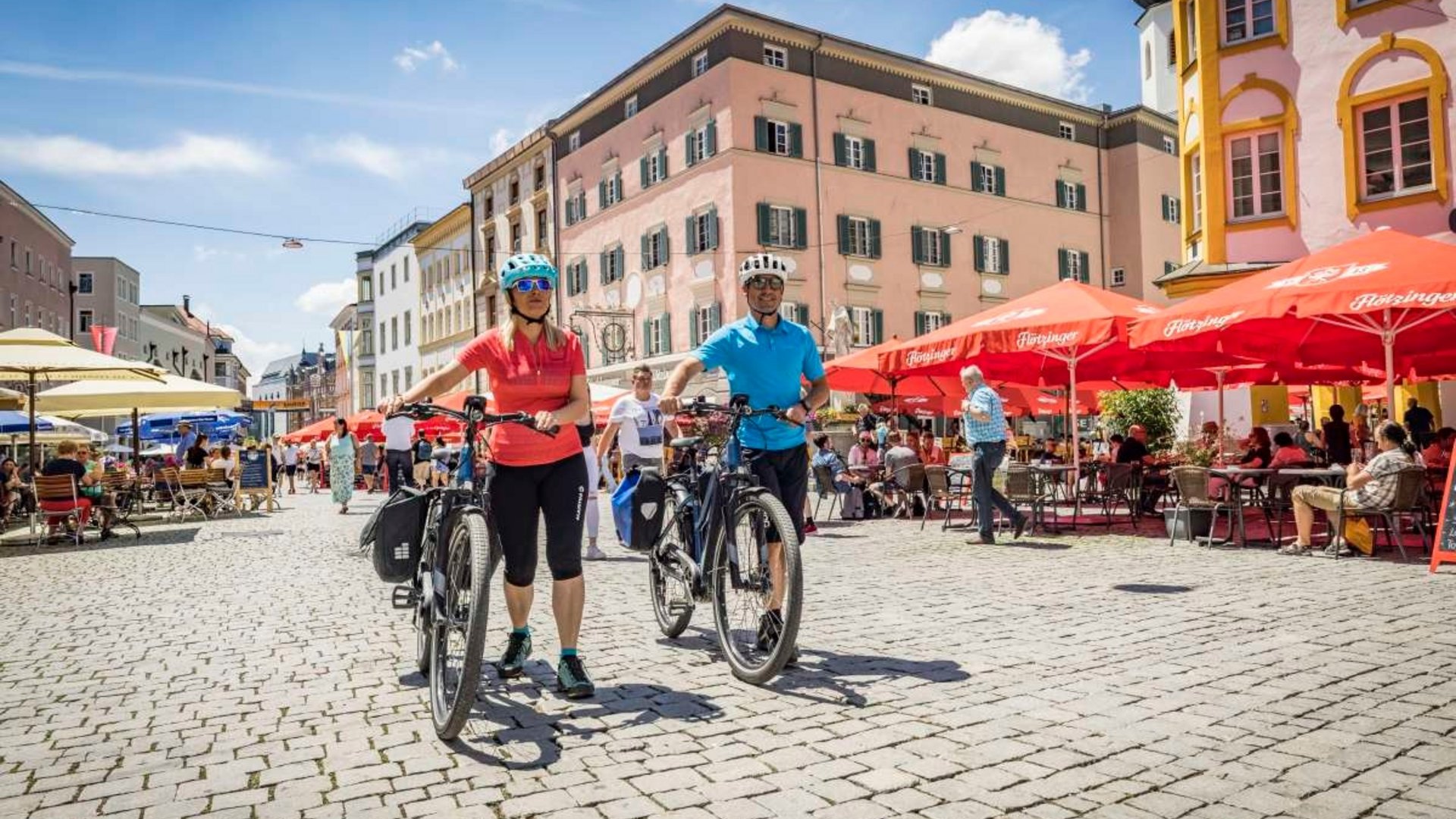 Two cyclists in a busy old town square with cafes and cobblestone