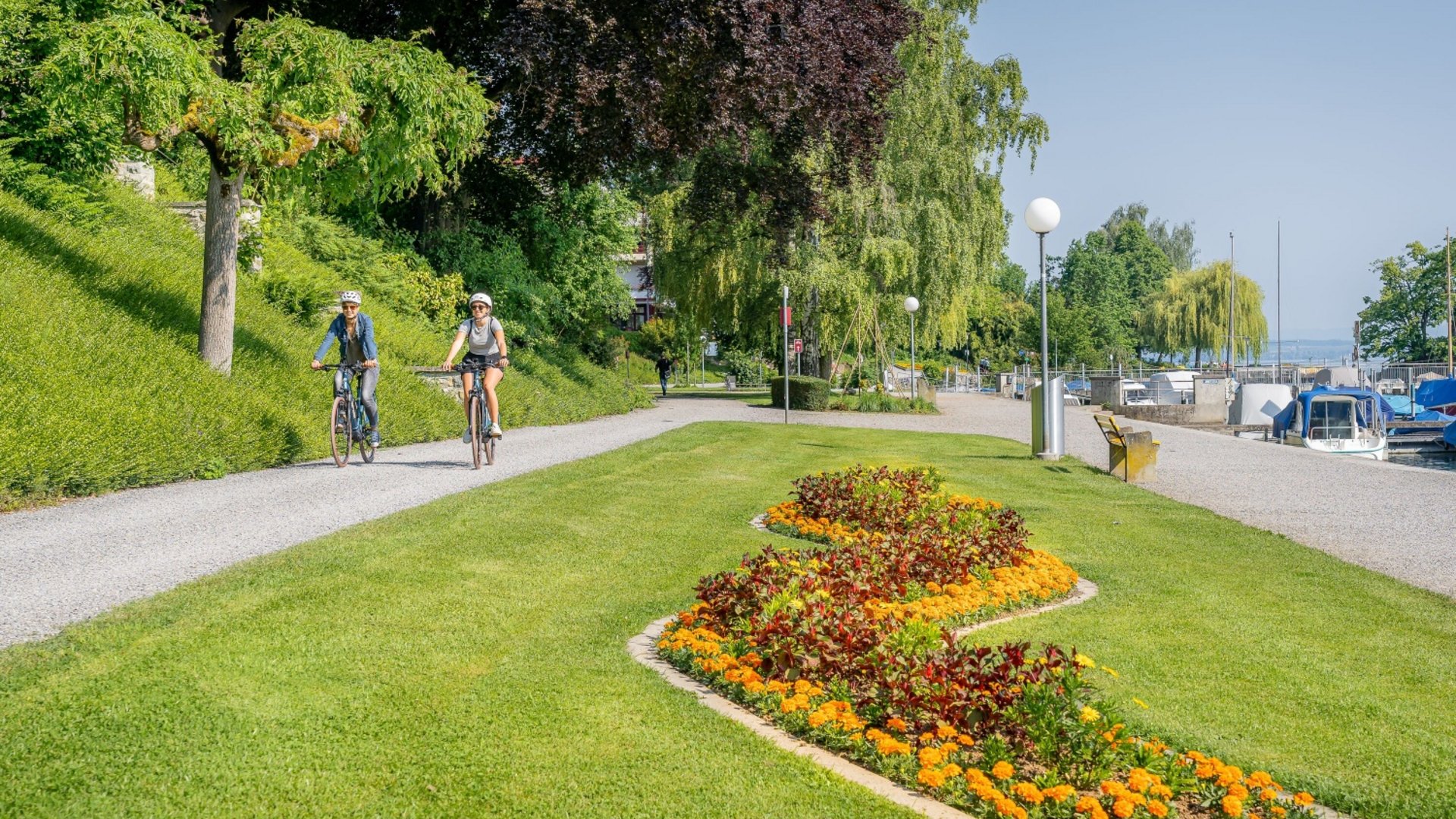 Two cyclists on a path beside flowerbed and water with boats